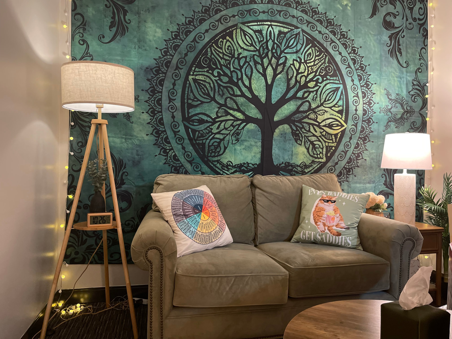 Cassidy's therapy office: an olive-green loveseat with playful pillows beneath a Tree of Life tapestry, framed by warm lamps and string lights